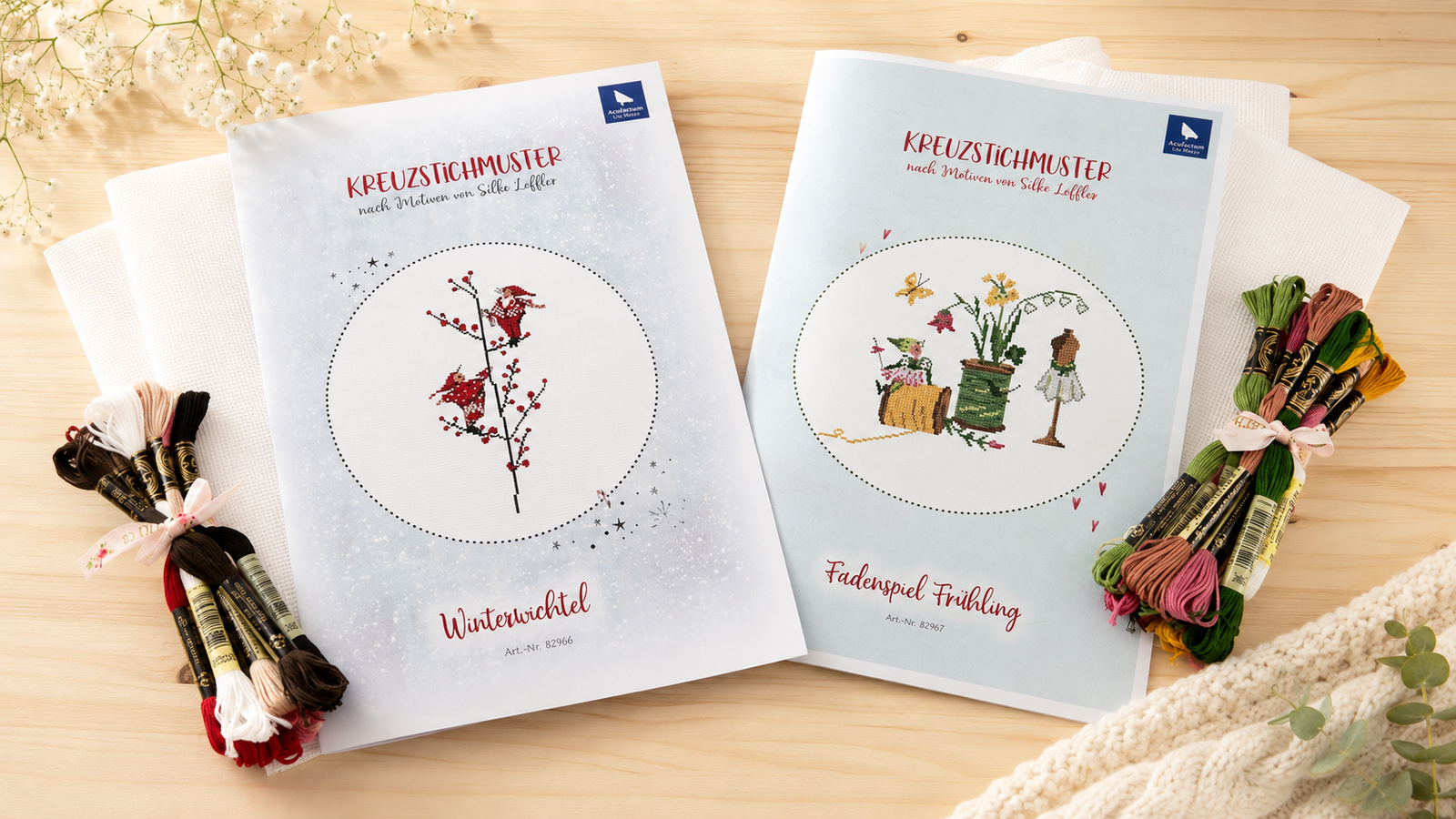 Acufactum cross stitch kits with Winterwichtel and Fadenspiel Frühling patterns, Aida fabric and coordinating embroidery floss arranged on a light wood table.