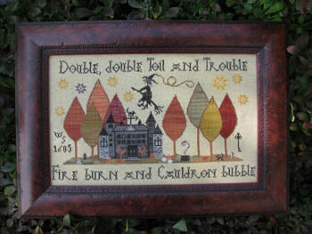 Framed cross-stitch art with witch, haunted house, and autumn trees, featuring Halloween-themed text.