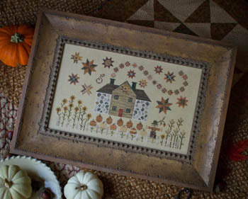 Framed autumn-themed cross-stitch art featuring a house, pumpkins, and stars in warm colors.