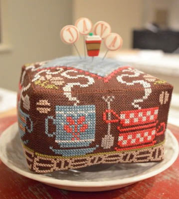 Colorful coffee-themed pincushion, square shape, featuring mugs and sewing motifs, made of fabric.