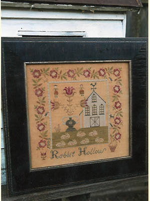 Framed Rabbit Hollow textile art with floral design, featuring a house and bunnies, in a dark wood frame.
