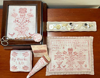 Vintage pink embroidered sampler set with decorative scissors, buttons, and accessories on a wooden table.
