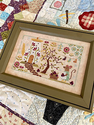 Framed embroidery art featuring a garden scene, colorful thread, stitched flowers, and house detail.