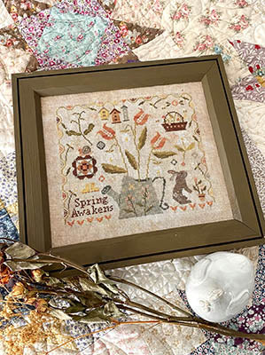 Framed cross-stitch art titled 'Spring Awakens', featuring colorful flowers, a bunny, and rustic home elements.