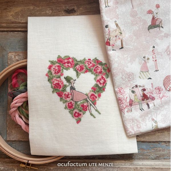 Pink floral heart cross-stitch design on linen fabric with sewing materials and patterned fabric.