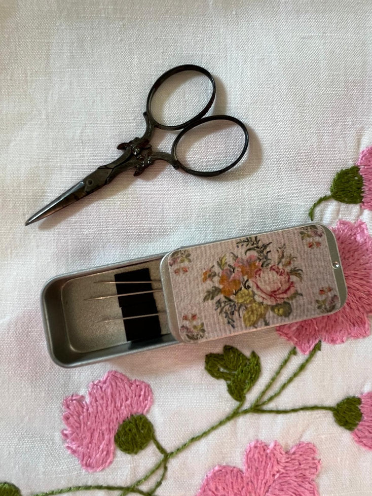Vintage floral sewing kit with scissors, needles in a decorative metal tin on embroidered fabric.