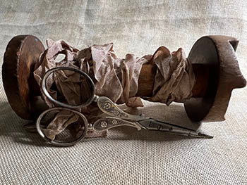Vintage wooden spool with tan fabric ribbon and ornate silver scissors on a textured surface.