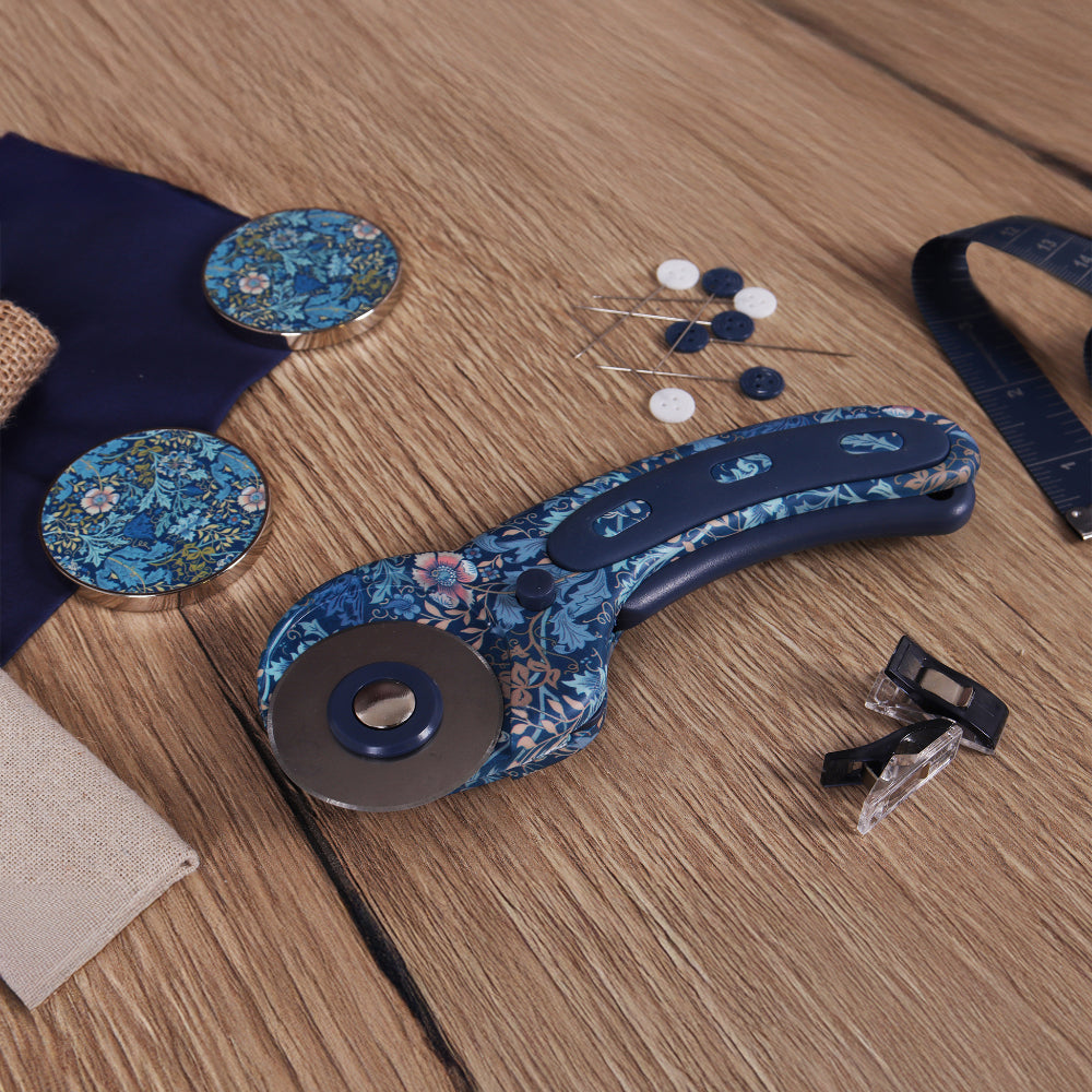 Floral-patterned rotary cutter with a blue handle on a wooden surface, alongside buttons and fabric.