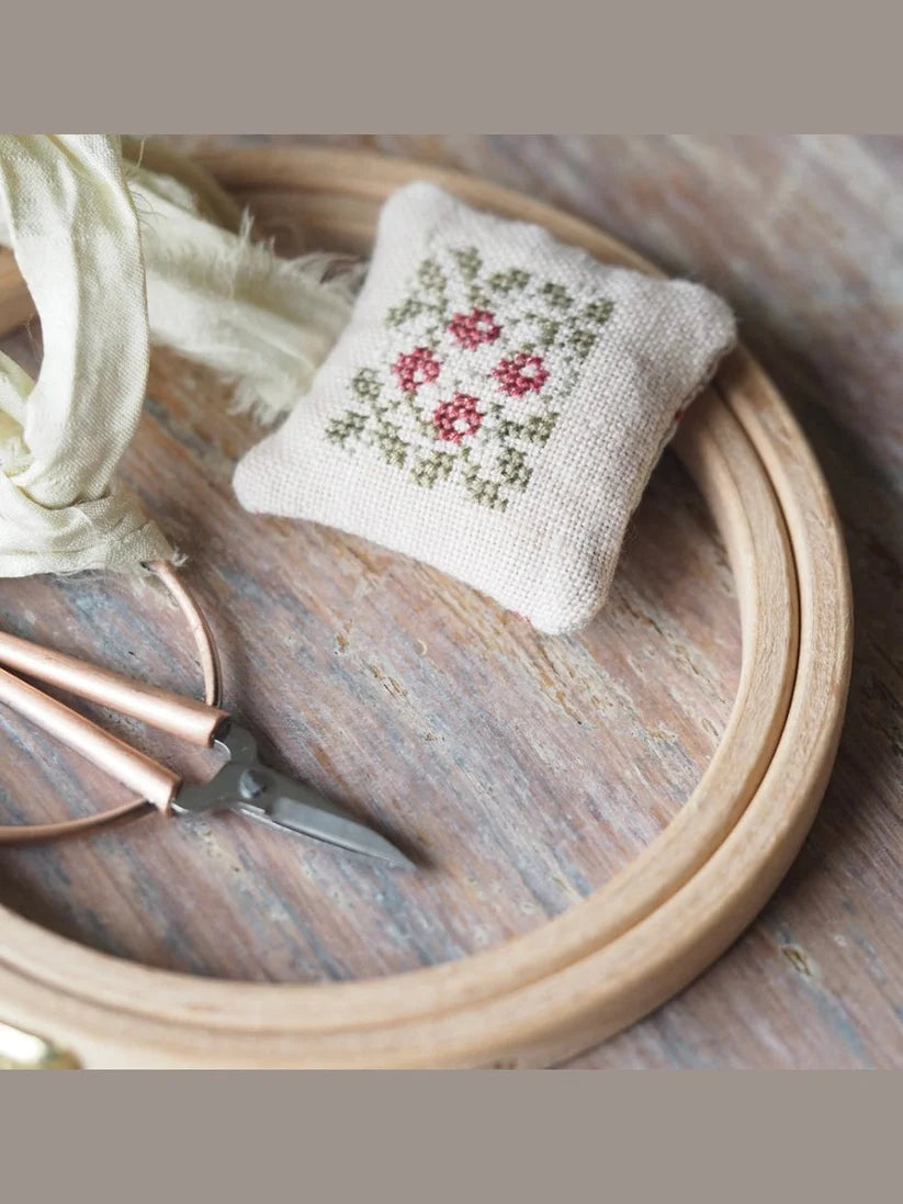 Handcrafted floral cross-stitch pincushion in cream linen with pink and green embroidery, scissors, and hoop.