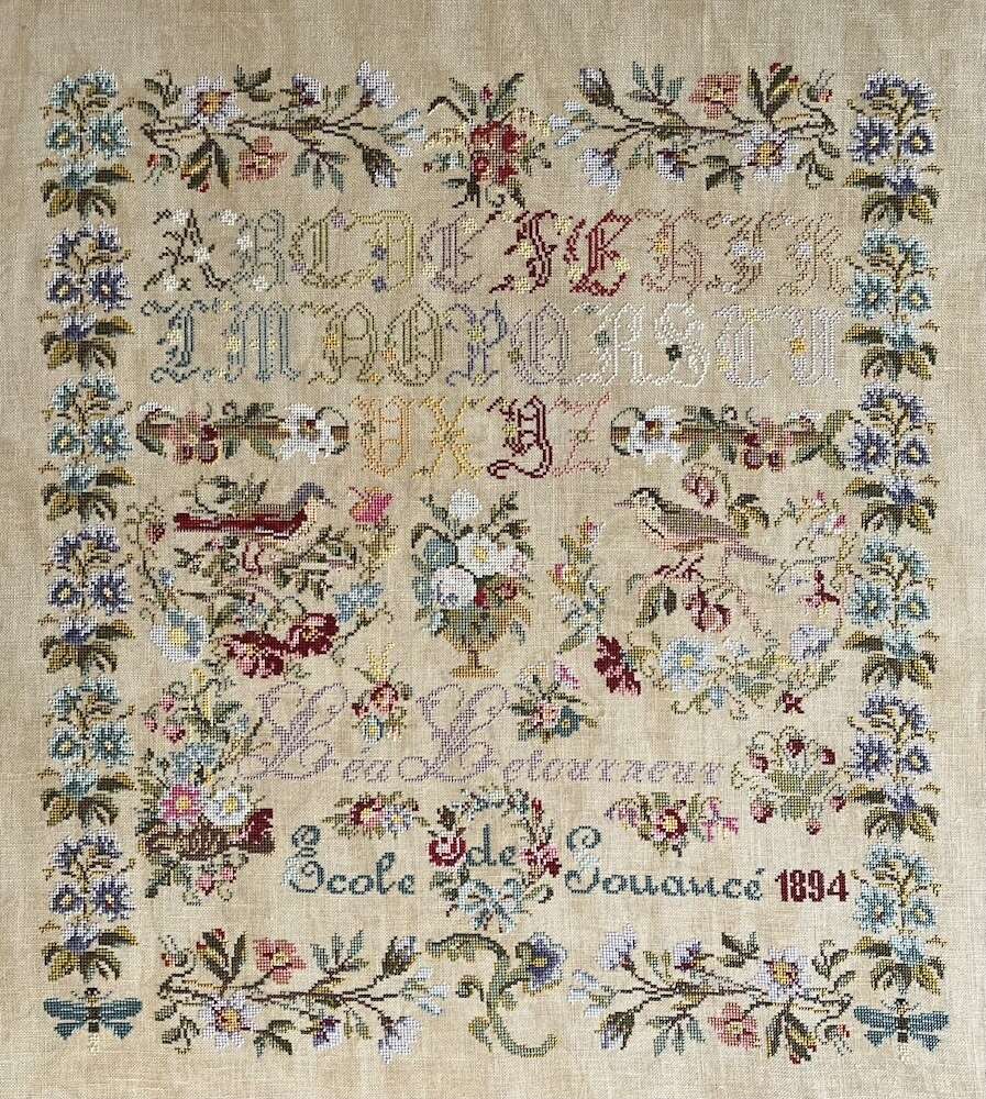 Vintage embroidery sampler with floral patterns and alphabet, featuring pastel colors on beige linen.