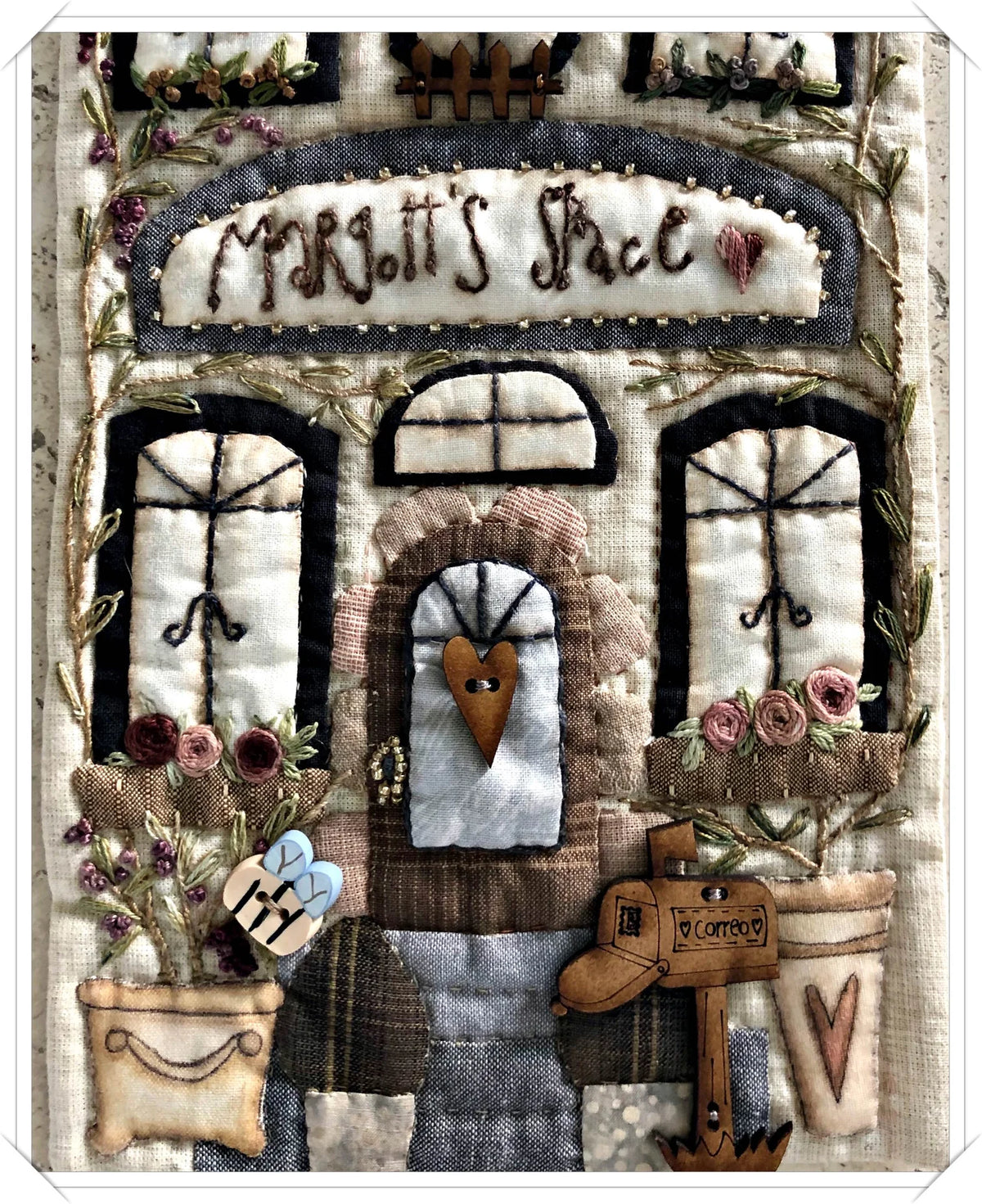 Hand-stitched fabric wall art featuring a cozy house with floral embellishments, heart-shaped details, and a mailbox.
