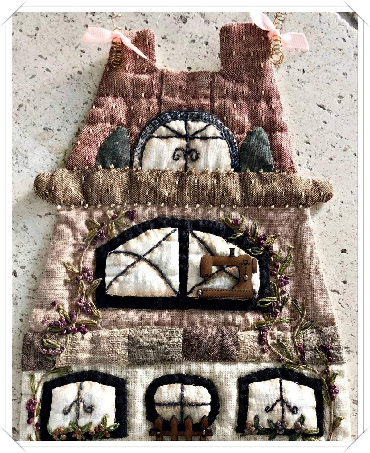 Quilted fabric wall hanging with sewing theme, featuring earthy tones, windows, and floral embroidery.
