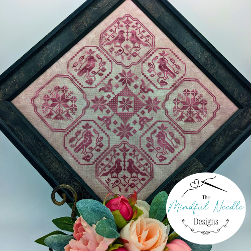 Framed cross-stitch art featuring pink patterns and snowflakes on light fabric, by The Mindful Needle Designs.