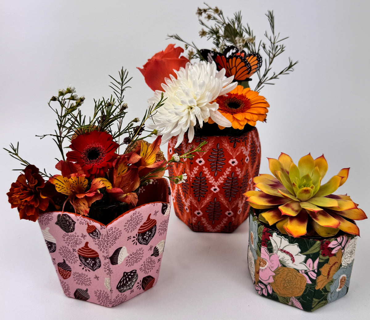 Three decorative flower pots: pink with shells, red with flowers, and green with floral design, showcasing vibrant blooms.