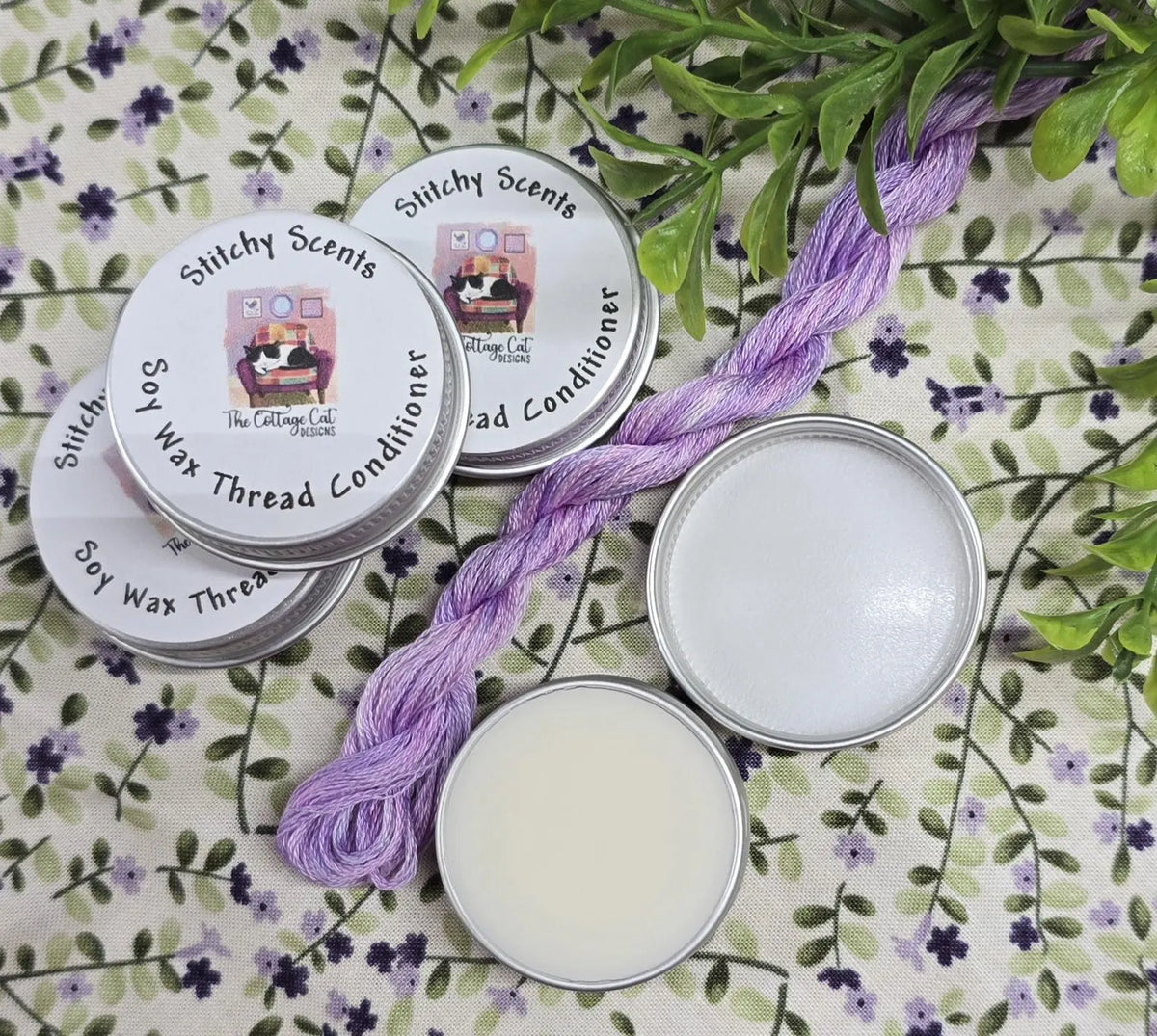 Stitchy Scents soy wax thread conditioner in silver tin, accompanied by lavender thread on floral background.