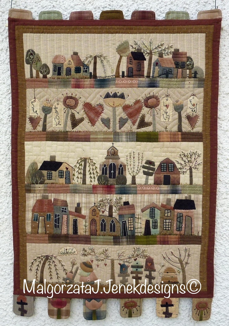 Quilted wall hanging featuring colorful houses, trees, and flowers in earthy tones, crafted from fabric.