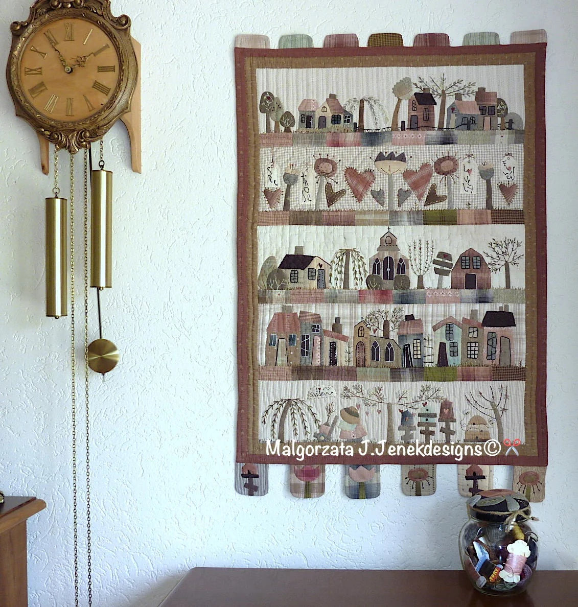 Colorful quilted wall hanging featuring whimsical houses, trees, and flowers in soft pastel tones.