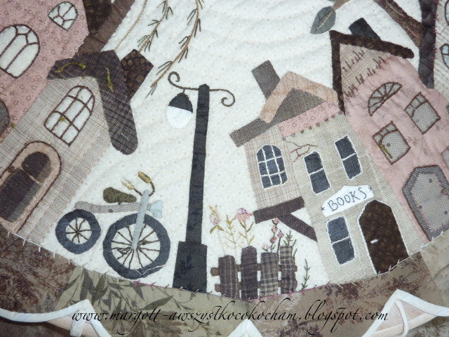 Quilt depicting a quaint village scene with houses, bicycle, lamppost, and decorative floral elements.