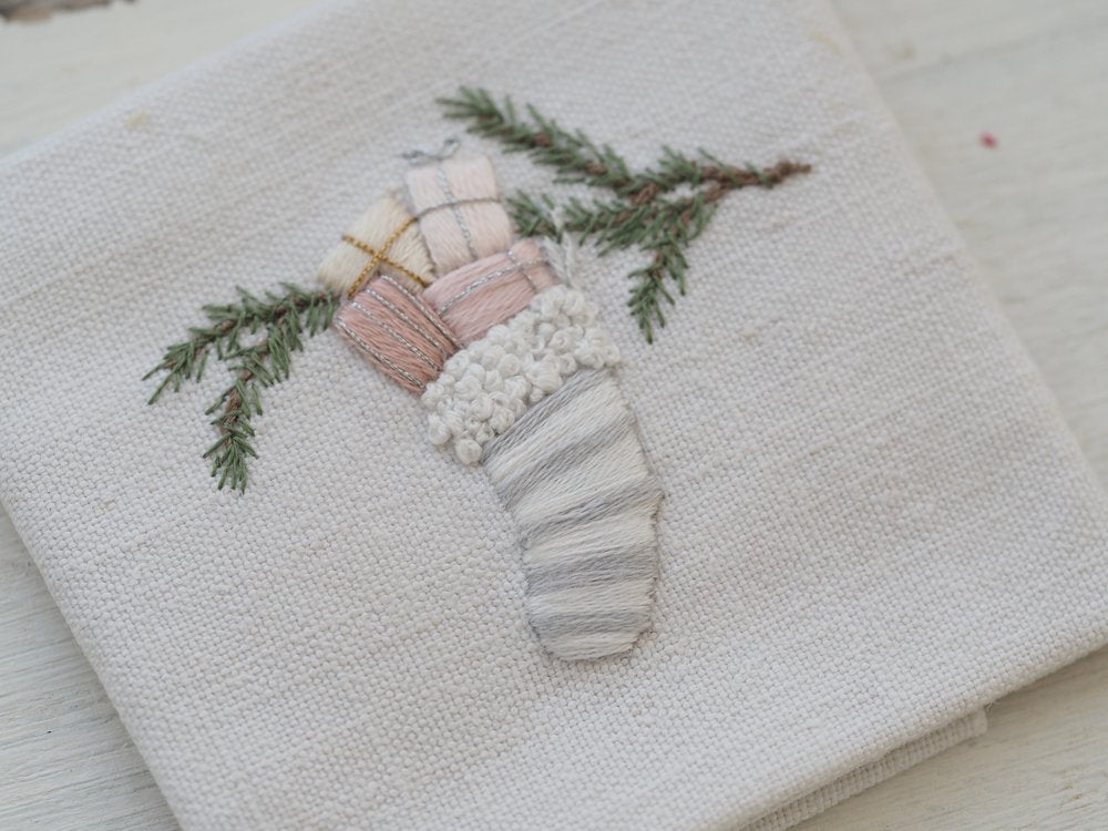 Hand-embroidered linen napkin with pastel gift boxes and green pine branches.